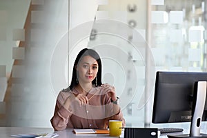 Attractive businesswoman holding pen and looking to camera while sitting in front of computer.