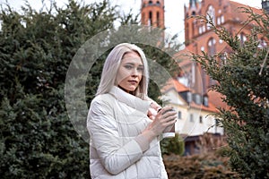 Attractive calm blond woman looking at camera, drinking cup of coffee, wearing white warm jacket. Travelling, journey.