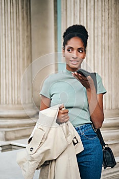 Attractive casual African Amerian girl with trench coat in hand dreamily talking on cellphone on street