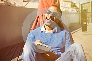 Attractive casually dressed young black male student with sunglasses making notes in copybook, preparing for lesson at