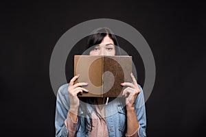 Attractive caucasian brunette girl in a shirt covering her face with a book and reading isolated on a black studio background.