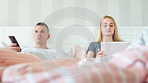 Attractive caucasian couple in bed. Man watching tv, woman browsing web on digital tablet. Couple in bed ignoring each other.