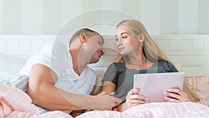 Attractive caucasian couple in bed. Woman holding digital tablet, man lovingly looking at her, hoping to catch her attention. Both