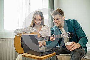 Attractive caucasian couple watch lessons of playing guitar using laptop