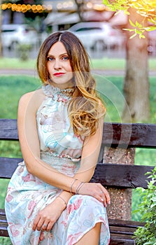 Attractive Caucasian girl sitting on a bench in the park. Light summer dress, floral print. Urban landscape.