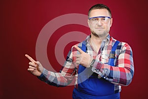 Attractive Caucasian male foreman in uniform and goggles points fingers at blank space. Foreman repairman maste studio