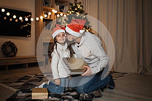 Attractive caucasian male sharing gift and kissing his cute preschool daughter in santa hat