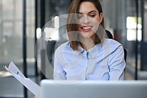 Attractive cheerful business woman checking paper documents in office, working on laptop