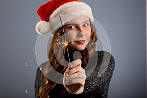 Attractive cheerful girl with curly hair in Christmas hat smiling, holding sparkler and drinking champagne, isolated on gray