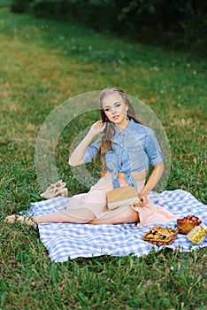 Attractive cheerful girl sitting on a blanket and spending the weekend relaxing in the park outdoors