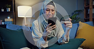 Attractive cheerful young Asia islam muslim beauty girl in hijab with casual smile sitting on couch use phone enjoy successful