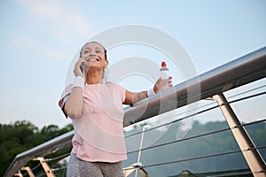 Attractive cheerful young Hispanic woman in pink t-shirt talking on mobile phone while resting on a city bridge after early