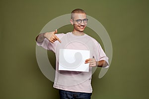 Attractive cheerful young man holding white empty paper board and smiling on green background