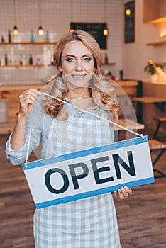 attractive coffee shop owner in apron holding sign open and smiling