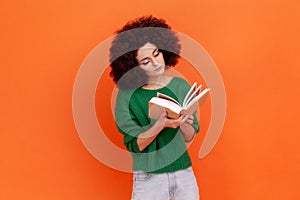 Attractive concentrated woman with Afro hairstyle wearing green casual style sweater reading interesting book, being very