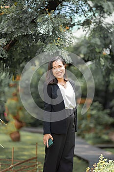 Attractive, Confidence young Asian Woman in business black suit is posting, smiling in front of leaves and tree with a relaxing