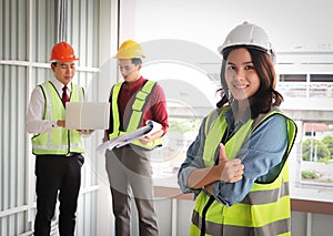 Attractive confident Asian female construction worker standing with arm crossed  and thumb up at construction site with two male