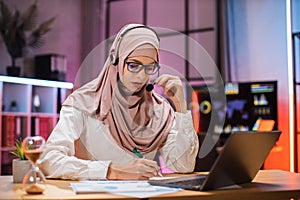 Attractive confident muslim business woman, office manager, wearing headset and hijab using laptop