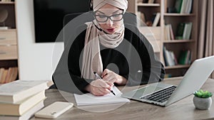 Attractive confident muslim business woman, office manager, wearing headset and hijab using laptop