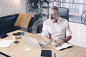 Attractive and confidental adult businessman using mobile laptop computer and making notes while working at the wooden