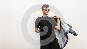 Attractive cool young man hipster puts on a denim jacket. Handsome stylish guy in a vintage T-shirt with trendy sunglasses posing