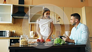 Attractive couple chatting in the kitchen early morning. Handsome man using tablet while his girlfriend cooking