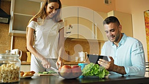 Attractive couple chatting in the kitchen early morning. Handsome man using tablet while his girlfriend cooking