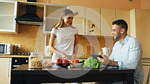 Attractive couple chatting in the kitchen early morning. Handsome man using tablet while his girlfriend cooking