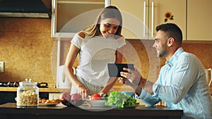 Attractive couple chatting in the kitchen early morning. Handsome man using tablet while his girlfriend cooking