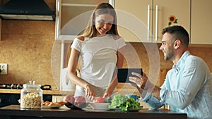 Attractive couple chatting in the kitchen early morning. Handsome man using tablet while his girlfriend cooking