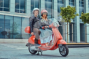 An attractive couple, a handsome man and female riding together on a red retro scooter in a city.