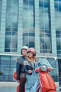 An attractive couple, a handsome man and female riding together on a red retro scooter in a city.