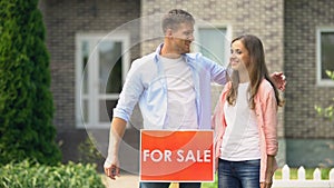 Attractive couple hugging installing for sale signboard in front of house, life