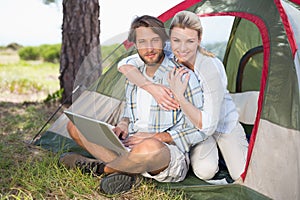 Attractive couple sitting by their tent using laptop smiling at camera
