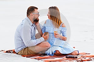 Attractive couple sitting together, looking each other, happy couple enjoying picnic on the white sand beach and drinking wine or