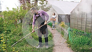 Attractive couple work near greenhouse. Man gardener in apron collect garbage in garden while his wife holding box with