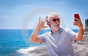 Attractive crazy senior man taking selfie at the sea, wearing red headphones and sunglasses. Carefree retired having fun using