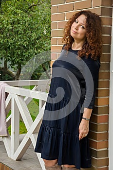 Attractive curly-haired red-haired woman for forty years standing on the porch of a country house in a blue dress with ruches.