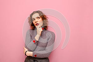 Attractive curly lady in casual clothes isolated on pastel pink background, looks away with pensive face on copy space. Pensive