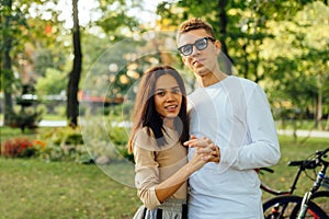 Attractive cute couple is standing in the park, posing for the camera, holding their hands and smiling. A caucasian man and