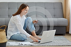 Attractive dark haired female wearing white shirt and jeans holding credit card in hands, using portable computer for entering