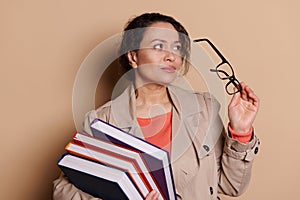 Attractive middle-aged female teacher holding eyeglasses, looking away, posing with stack of books, isolated background