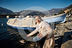 Attractive delightful young woman in beige coat, sitting nears boats by the Como lake, relaxing in the lakefront photo