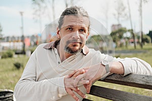 An attractive elderly 50-year-old man with long hair in the tail sits in a public garden on a bench. Warm summer day