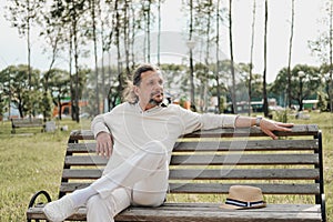 An attractive elderly 50-year-old man with long hair in the tail sits in a public garden on a bench. Warm summer day