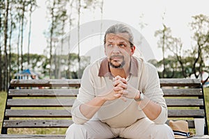 An attractive elderly 50-year-old man with long hair in the tail sits in a public garden on a bench. Warm summer day