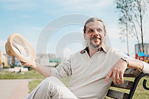 An attractive elderly 50-year-old man with long hair in the tail sits in a public garden on a bench. Warm summer day