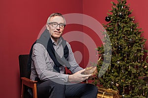Attractive elderly man holds Christmas presents gold box against red studio wall background and green decorated Christmas tree