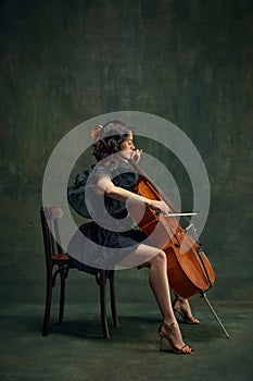 Attractive, elegant young woman, cellist, musician in black dress sitting on chair and playing cello against dark