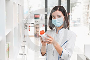 Attractive ethnicity female pharmacist wearing medicine mask holding a pill container with antiviral medicine